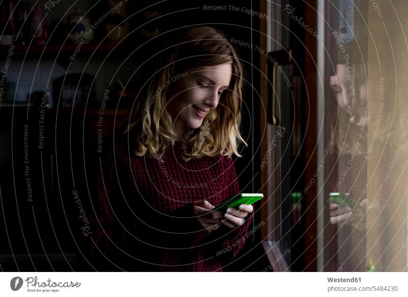 Lächelnde Frau steht zu Hause am Fenster und schaut auf ihr Smartphone iPhone Smartphones weiblich Frauen Handy Mobiltelefon Handies Handys Mobiltelefone