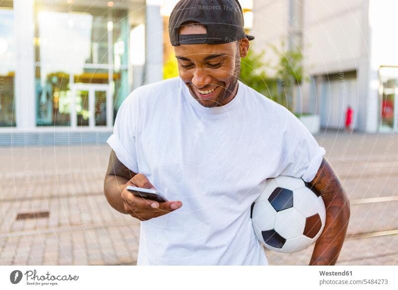 Lachender junger Mann mit Fussball, der auf sein Handy schaut Smartphone iPhone Smartphones Fußball Fußbälle Männer männlich Mobiltelefon Handies Handys