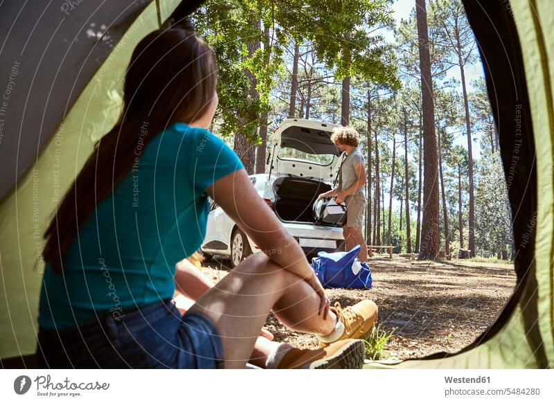 Junge Frau sitzt in einem Zelt und sieht zu, wie ihr Freund das Auto packt zusehen zusehend Zelte beladen einpacken einladen Wagen PKWs Automobil Autos Paar