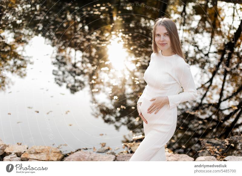 Mütter Tag. Schöne junge schwangere Frau in weißen engen Strickkleid mit kleinen Bauch entspannen im Freien durch den Teich mit Reflexion Natur und Sonne im Park und Blick auf die Kamera. Frauen Tag