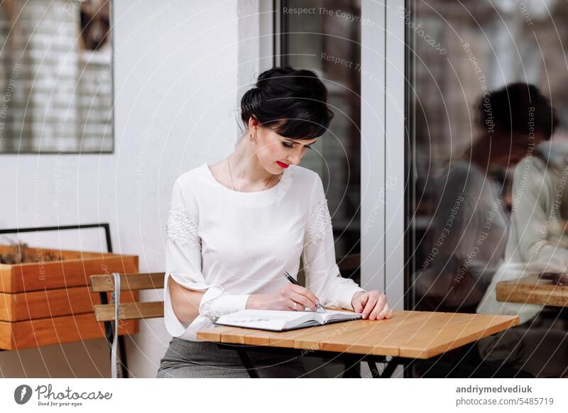 Serious fokussiert intelligente Geschäftsfrau schreibt Notizen auf Notizblock, machen Tagesordnung auf persönlichen Organizer. Frau in Business-Anzug sitzt am Tisch im Café auf der Terrasse, Arbeit und Event-Planung.