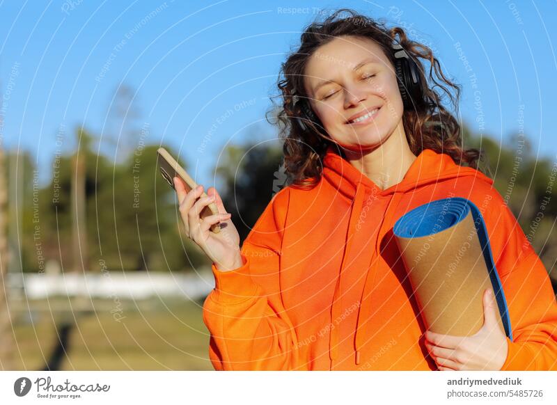 Glückliche junge Frau mit Kopfhörern und Yogamatte, hört Musik auf dem Smartphone im Freien. Lächelndes Mädchen mit geschlossenen Augen in lebhaften orangefarbenen Sweatshirt genießt Lieblings-Playlist nach dem Training in grünen Park