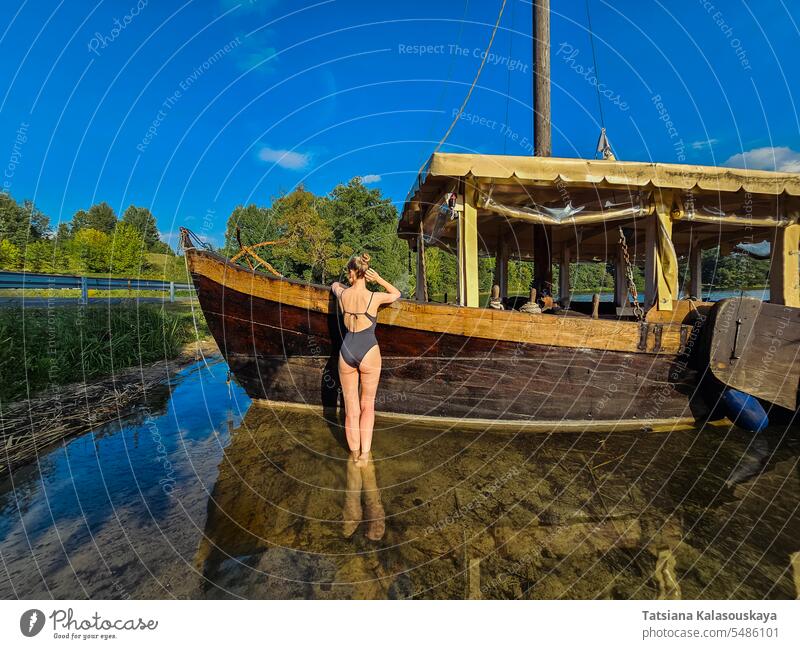 Eine Frau ruht sich im See aus und steht in der Nähe eines alten Holzschiffs. Attraktive fit Frau ruht in der Nähe des Sees im Sommer. schlank Badeanzug Stehen