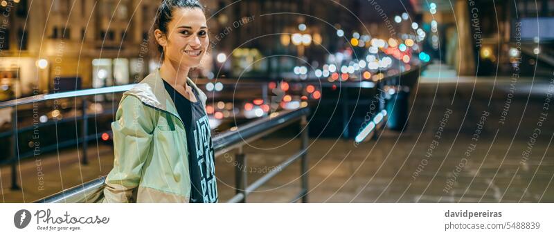 Frau in Sportkleidung blickt in die Kamera mit einer nächtlichen Stadt im Hintergrund Läufer tragend Sportbekleidung Porträt Blick Fotokamera Glück Lächeln