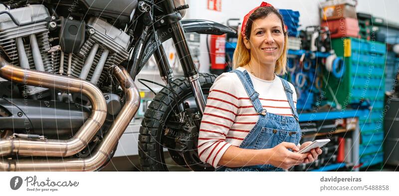 Mechanikerin mit Telefon in der Hand, sitzend auf einer Plattform mit Motorrad in der Fabrik Porträt Frau Lächeln Fröhlichkeit Beteiligung benutzend Funktelefon
