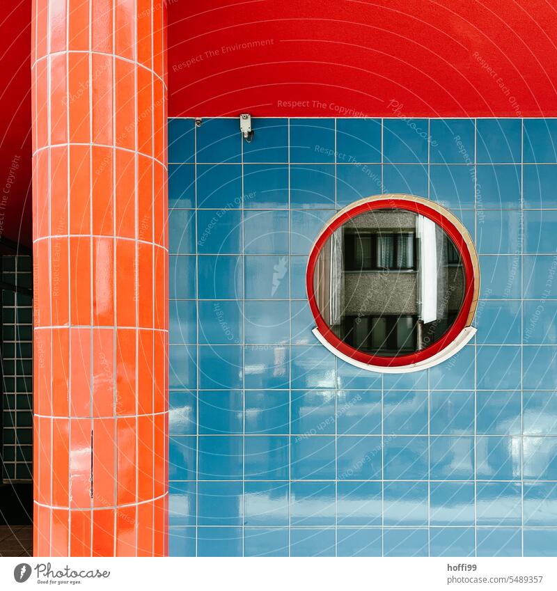 Farbenfroh in den Tag - Fassade mit blau, rot, orangen Fliesen und rundem Fenster in dem sich eine graue Fassade, sowie Fotograf und ein paar Autos, spiegelt.