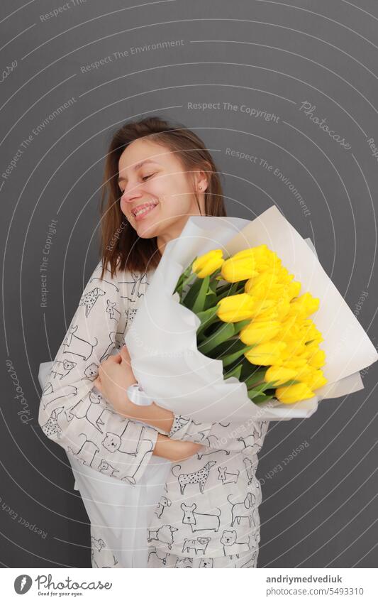 Sehr schöne junge glückliche Frau hält großen und schönen blühenden Strauß frischer gelber Tulpen Blumen auf der grauen Wand Hintergrund. Internationaler Frauentag
