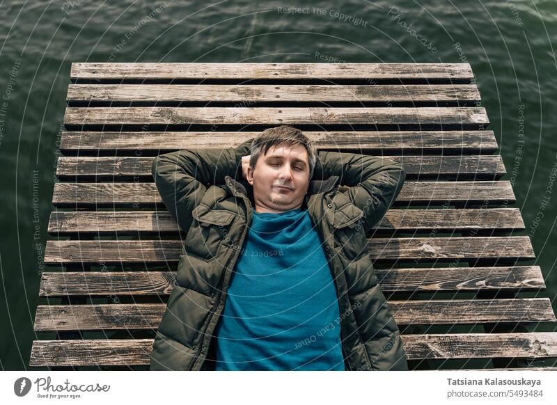Ein Mann liegt in Jacke und Pullover mit geschlossenen Augen auf einem Holzsteg am See. Trägerkleid Lügen entspannend hölzern Bootsanlegestelle Dock genießend