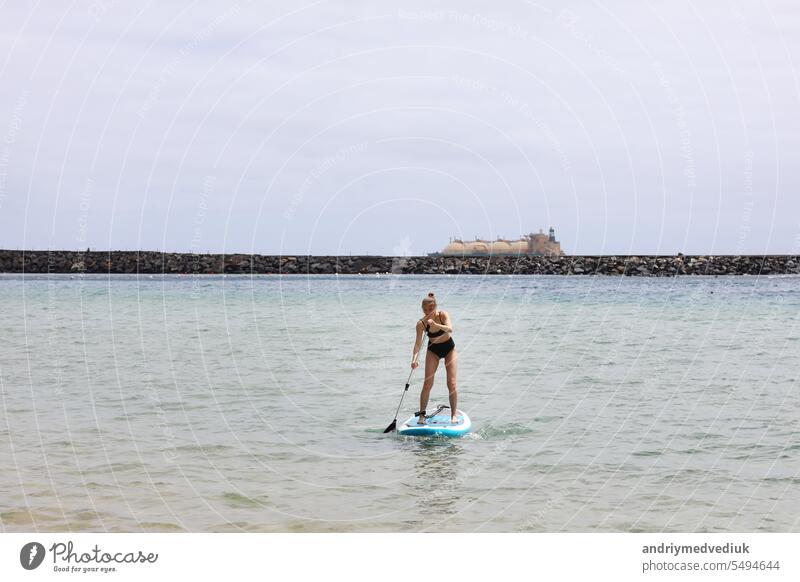 Junge Frau in stilvollen schwarzen Badeanzug mit einem Paddel anmutig Balancieren auf einem SUP-Board, Gleiten entlang der ruhigen Ozean auf sonnigen Sommertag. Gesunder Lebensstil und extreme Erholung Konzept.