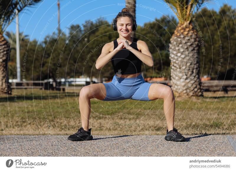 Junge gesunde Frau in Sportkleidung hört Musik über Kopfhörer, während sie Fitness-Beinübungen im Freien im Park mit Palmen macht. Workout zur Stärkung der Beckenbodenmuskulatur im Sommerurlaub