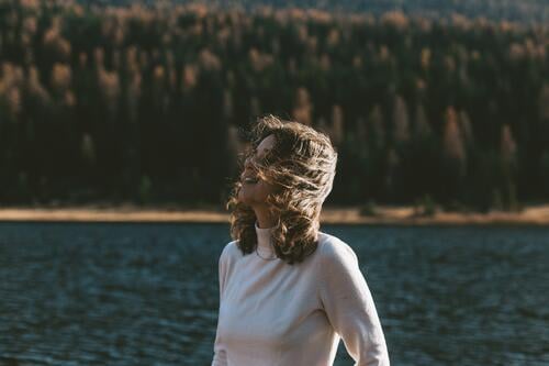 eine Frau steht vor einem herbstlichen Wald mit dem Wind in ihren Haaren Behaarung windig windiges Wetter fallen Herbst authentisch friedlich Himmel Wolken