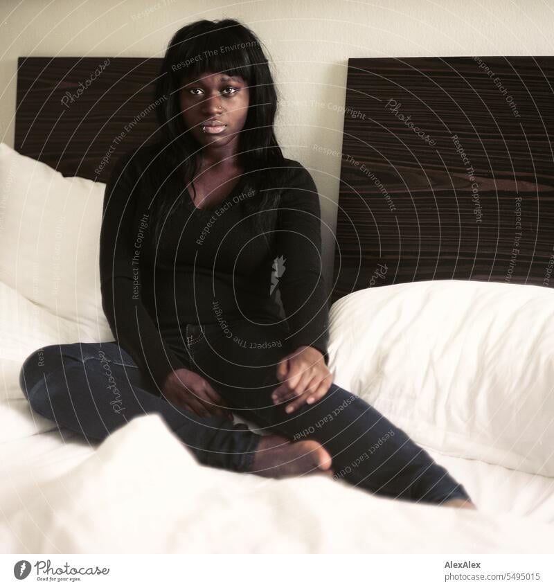 analoges Portrait einer Frau mit langen, schwarzen Haaren und schwarzem Pullover und Jeans auf einem Bett in einem Hotelzimmer junge Frau schön Lippen