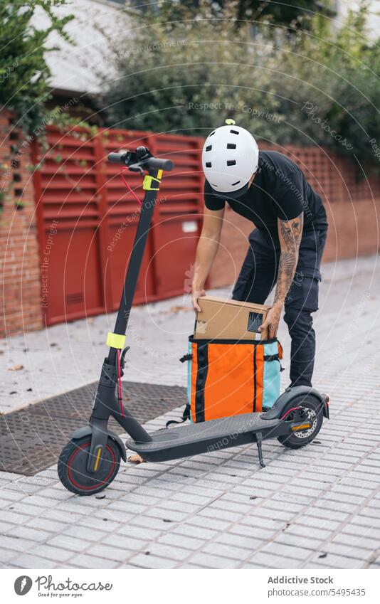 Männlicher Kurier mit Kiste in der Nähe eines elektrischen Kick-Scooters Auslieferer entfernen Tasche Kickroller Verkehr umweltfreundlich Versand Straßenbelag