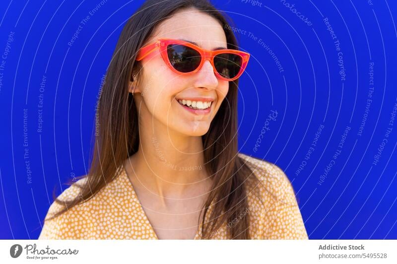 Lächelnde Frau mit Sonnenbrille vor blauem Hintergrund trendy Stil hell positiv Glück heiter Freizeit jung Outfit modern Vorschein Brille Freude Accessoire