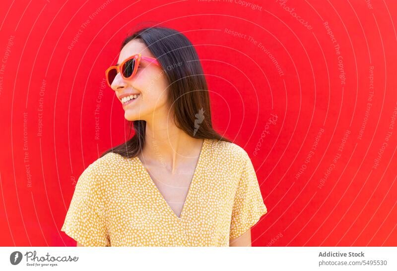 Lächelnde Frau mit Sonnenbrille vor rotem Hintergrund trendy Stil hell positiv Glück heiter Freizeit jung Outfit modern Vorschein Brille Freude Accessoire