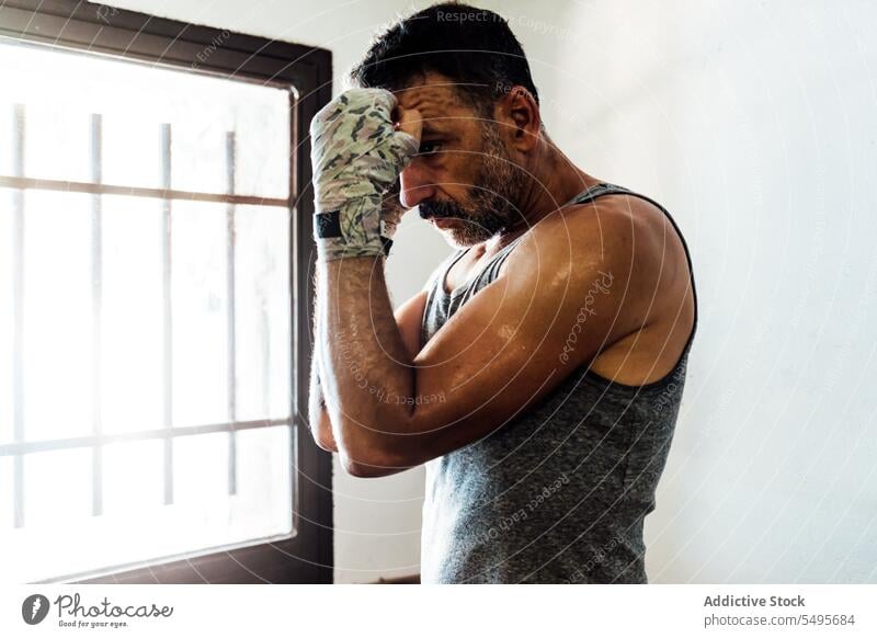 Kräftiger erwachsener Mann, der mit in ein Tuch eingewickelten Händen in der Nähe eines Fensters in einem Raum steht muskulös Training Athlet Boxer Sport