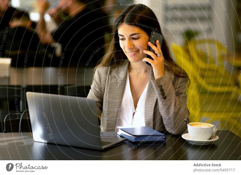 Lächelnde Frau, die mit ihrem Smartphone spricht, während sie in einem Café auf ihrem Laptop surft Telefonanruf reden freiberuflich abgelegen Arbeit Gerät