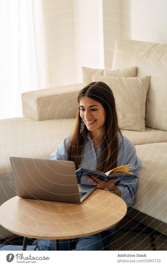 Glückliche junge Frau liest ein Buch, während sie mit einem Laptop in der Nähe des Sofas sitzt lesen positiv Freizeit Hobby Lächeln Komfort zu Hause Gerät