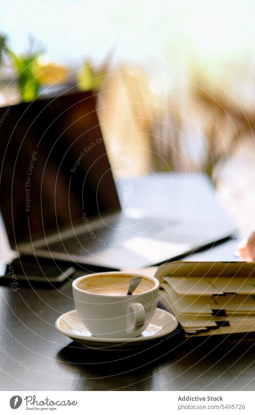 Tasse Kaffee auf dem Tisch und Buch mit Laptop auf dem Tisch Cappuccino Heißgetränk Löffel Notebook Café Smartphone Arbeitsbereich Morgen modern Kantine Planer