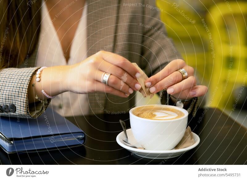 Crop-Frau, die Zucker in eine Tasse Kaffee im Café gibt hinzufügen Heißgetränk Tagebuch Buch Planer Veranstalter Tisch lesen jung Cappuccino formal Kantine