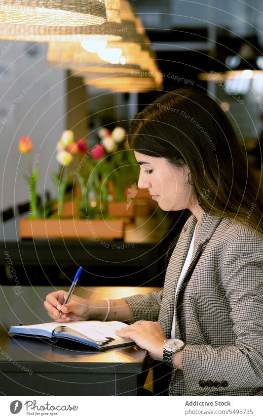 Konzentrierte junge Frau sitzt am Tisch und macht Notizen in einem Café zur Kenntnis nehmen schreiben Notebook Lächeln Notizblock Planer Schreibstift positiv