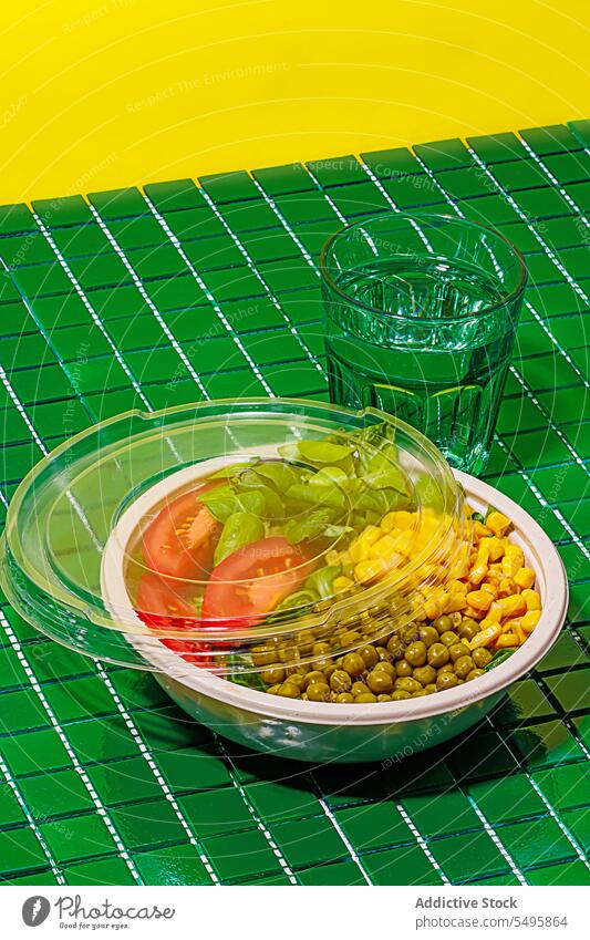 Salatschüssel auf grünem Hintergrund mit Plastikabdeckung neben einem Glas Wasser Schalen & Schüsseln Salatbeilage Lebensmittel Scheibe Tomate Spinat Blatt Mais
