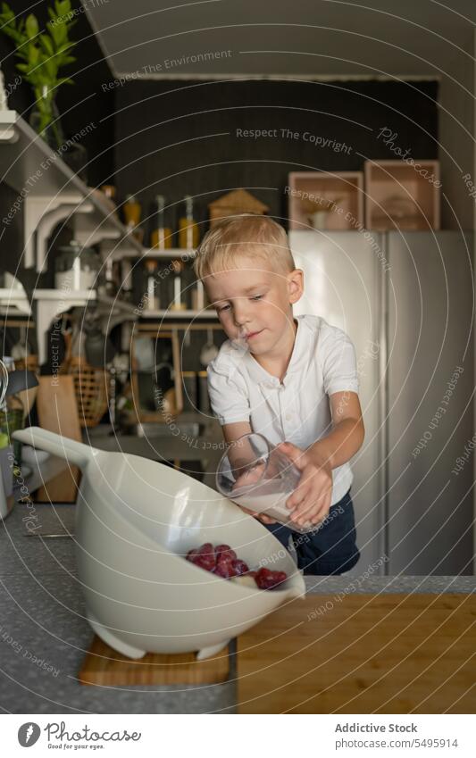 Junge gießt Milch in eine Schüssel mit Erdbeeren in der Küche melken Glas Gießen Essen zubereiten Kind Frucht heimwärts niedlich blondes Haar frisch