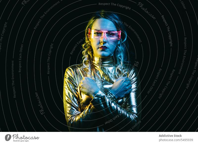 Kybernetisches junges Mädchen mit verschränkten Armen in geschlossenen Fäusten vor dunklem Hintergrund kybernetisch lockig Behaarung blond Brille futuristisch