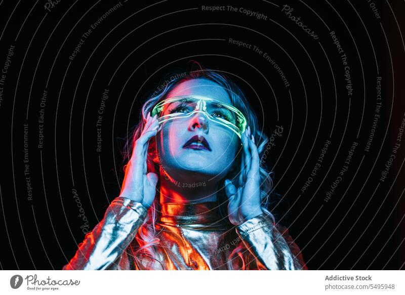 Kybernetisches junges Mädchen vor dunklem Hintergrund kybernetisch lockig Behaarung blond Brille futuristisch anhaben hell reflektierend Bekleidung emotionslos
