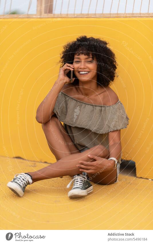 Glückliche Frau, die über ihr Smartphone spricht, während sie auf einer gelben Fläche sitzt Telefonanruf Funktelefon reden benutzend sprechen Gespräch Lächeln