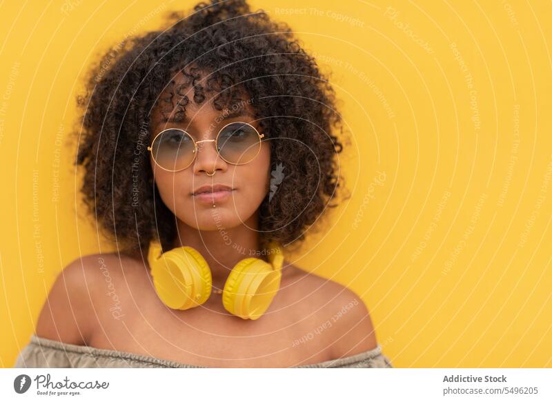 Junge Frau mit Kopfhörern und Brille schaut in die Kamera vor gelbem Hintergrund Piercing Sonnenbrille krause Haare Model Stil modern trendy Porträt Accessoire
