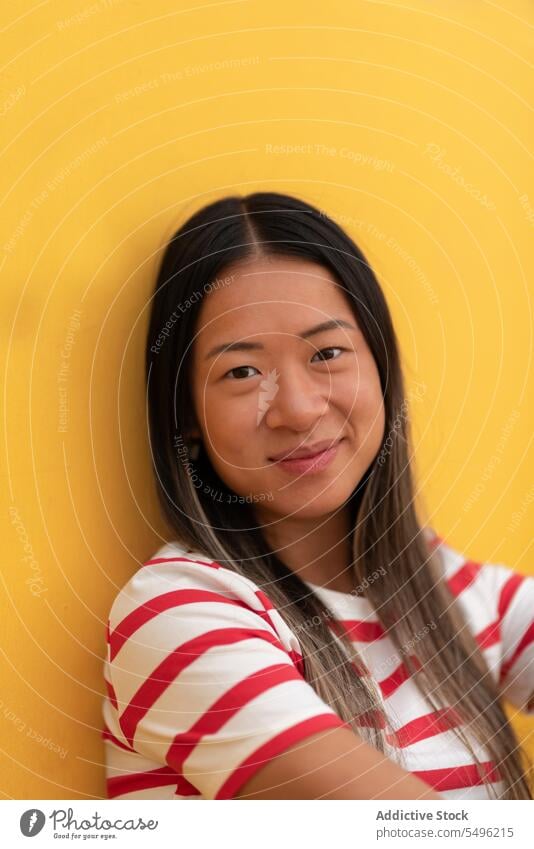 Lächelnde Frau mit Brille sitzt an einer gelben Wand positiv Glück sitzen lässig Streifen Porträt froh Optimist Mode Vergnügen angenehm asiatisch selbstbewusst