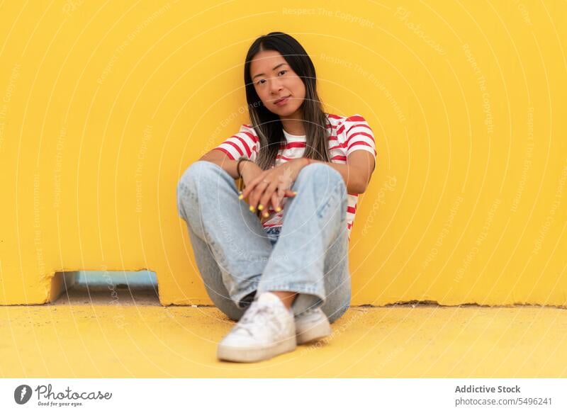 Asiatische Frau sitzt auf dem Boden vor gelbem Hintergrund lässig selbstbewusst fettarm Windstille sitzen trendy Outfit positiv Persönlichkeit allein