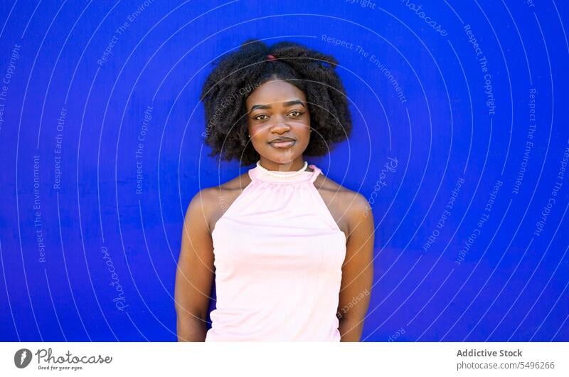 Lächelndes schwarzes Teenager-Mädchen im Freizeitkleid auf blauem Hintergrund Porträt Afro-Look Frisur Vorschein Make-up krause Haare Windstille lässig jung