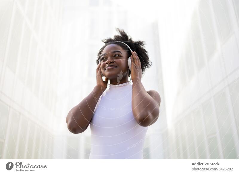 Fröhliches schwarzes Teenager-Mädchen, das Kopfhörer berührt zuhören Musik Straße Gesang genießen Afro-Look positiv Apparatur Gerät Lächeln Wiedergabeliste Frau