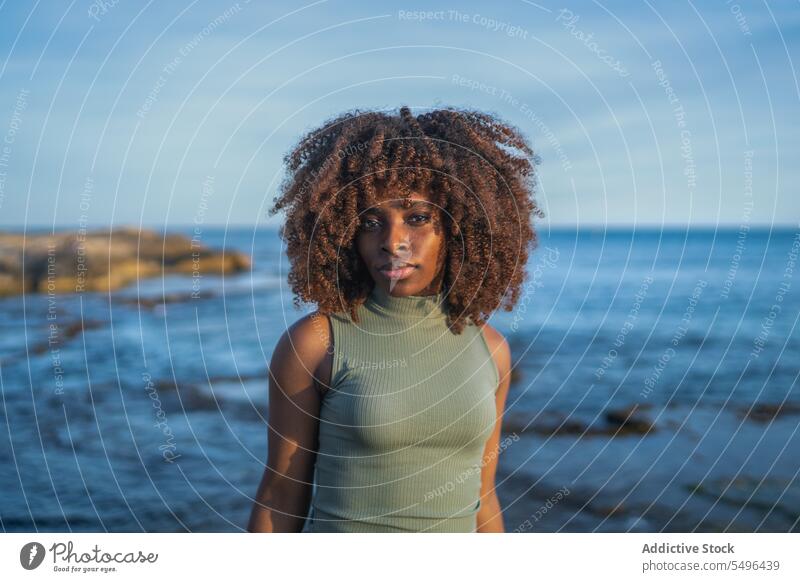 Ernste afroamerikanische junge Frau, die in die Kamera schaut, während sie vor einem verschwommenen Strand steht schwarz Dame ernst besinnlich nachdenklich