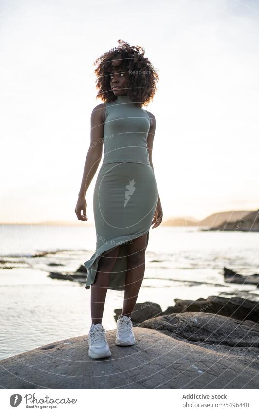 Ernste afroamerikanische junge Frau, die wegschaut, während sie vor einem verschwommenen Strand bei Sonnenuntergang steht schwarz Dame ernst besinnlich