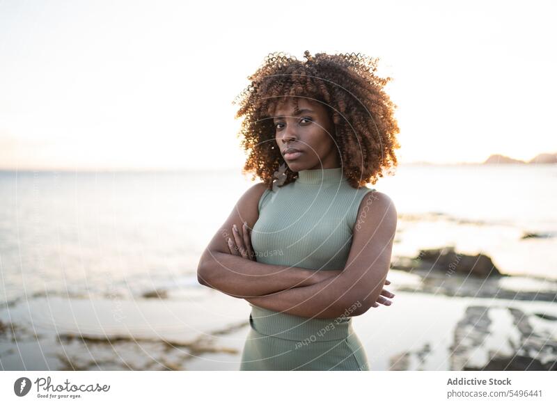 Ernste afroamerikanische junge Frau im Kleid schaut in die Kamera, während sie vor einem verschwommenen Strand bei Sonnenuntergang steht schwarz Dame