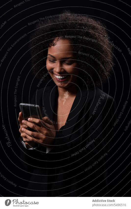 Lächelnde ethnische Frau mit Smartphone in schwarzem Studio benutzend Browsen Telefon Glück positiv online Nachricht Mobile hispanisch jung Apparatur Gerät