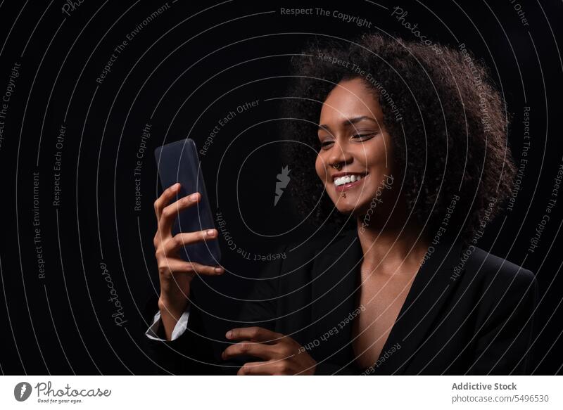 Lächelnde ethnische Frau mit Smartphone in schwarzem Studio benutzend Browsen Telefon Glück positiv online Nachricht Mobile hispanisch jung Apparatur Gerät