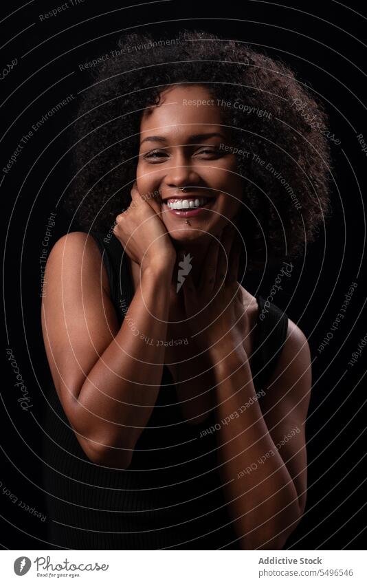 Fröhliche ethnische Frau berührt Wange in dunklem Studio positiv Gesicht berühren heiter Lächeln Porträt Vorschein Freizeit sorgenfrei Glück Afro-Look