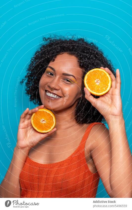 Fröhliche ethnische Frau mit Orangenscheiben in blauem Studio Lächeln Scheibe orange Frucht Hautpflege Schönheit Glück Porträt positiv krause Haare jung Sommer