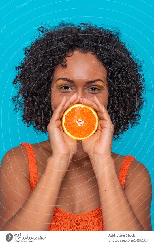 Glückliche ethnische Frau, die den Mund mit einer Orangenscheibe bedeckt Lächeln orange Zitrusfrüchte Frucht Scheibe Hautpflege Porträt jung krause Haare