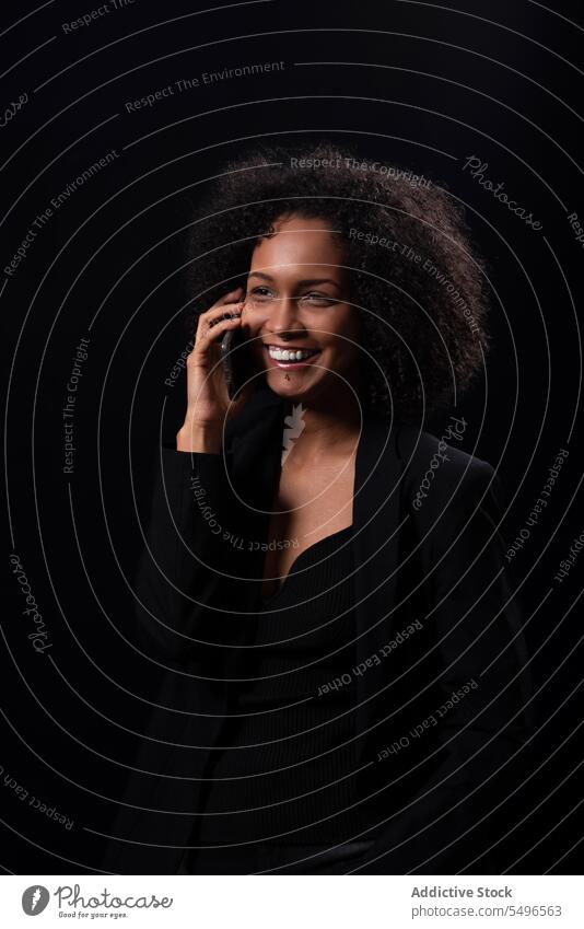 Lächelnde ethnische Frau mit Smartphone in schwarzem Studio benutzend Telefon Glück positiv online Mobile hispanisch jung Apparatur Gerät heiter