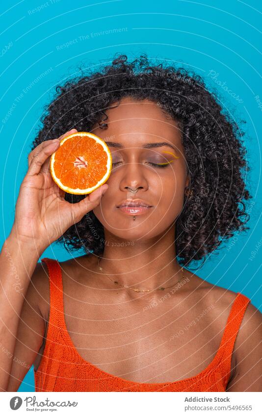 Ernste ethnische Frau bedeckt Auge mit Orangenscheibe orange Zitrusfrüchte Frucht Auge abdecken Scheibe Hautpflege Porträt jung krause Haare hispanisch hell