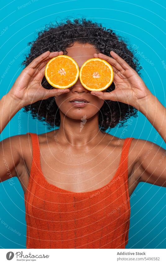 Ernste ethnische Frau, die ihre Augen mit Orangenscheiben in einem blauen Studio bedeckt orange Zitrusfrüchte Frucht Auge abdecken Scheibe Hautpflege Porträt
