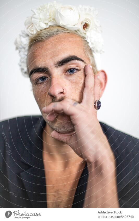 Selbstbewusster junger Mann mit blauen Augen und Blumenkrone in der Nähe einer weißen Wand im Zimmer Porträt Windstille Make-up Vorschein unrasiert Schnurrbart