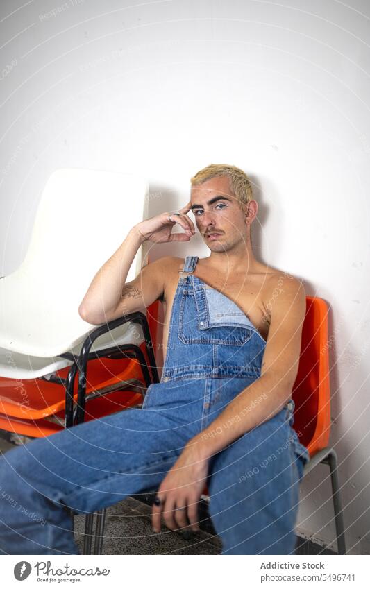 Junger Mann ohne Hemd mit nackten Schultern und in Jeans schaut in die Kamera, während er auf einem Stuhl sitzt Make-up Wand selbstbewusst nackte Schultern