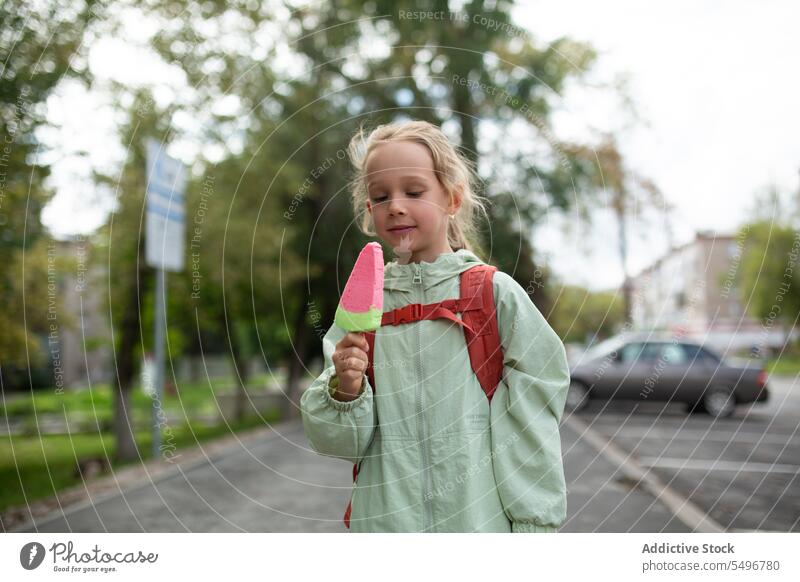 Glückliches Kind mit Rucksack auf dem Gehweg stehend und Eis genießend Speiseeis Straße Lächeln Bürgersteig heiter niedlich Kindheit PKW Mädchen bezaubernd