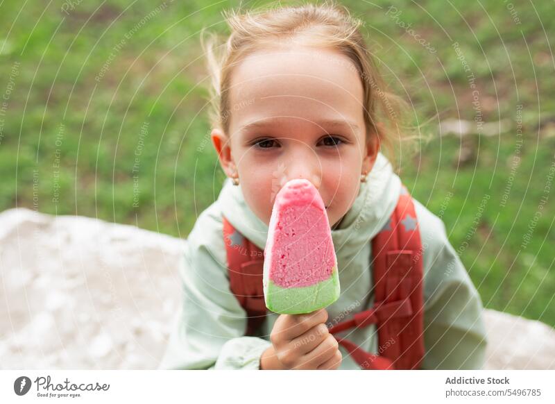 Glückliches Kind mit Rucksack steht in der Nähe von grasbewachsenen Feld und genießt Eiscreme Speiseeis Park Lächeln Weg heiter niedlich Kindheit grün Mädchen